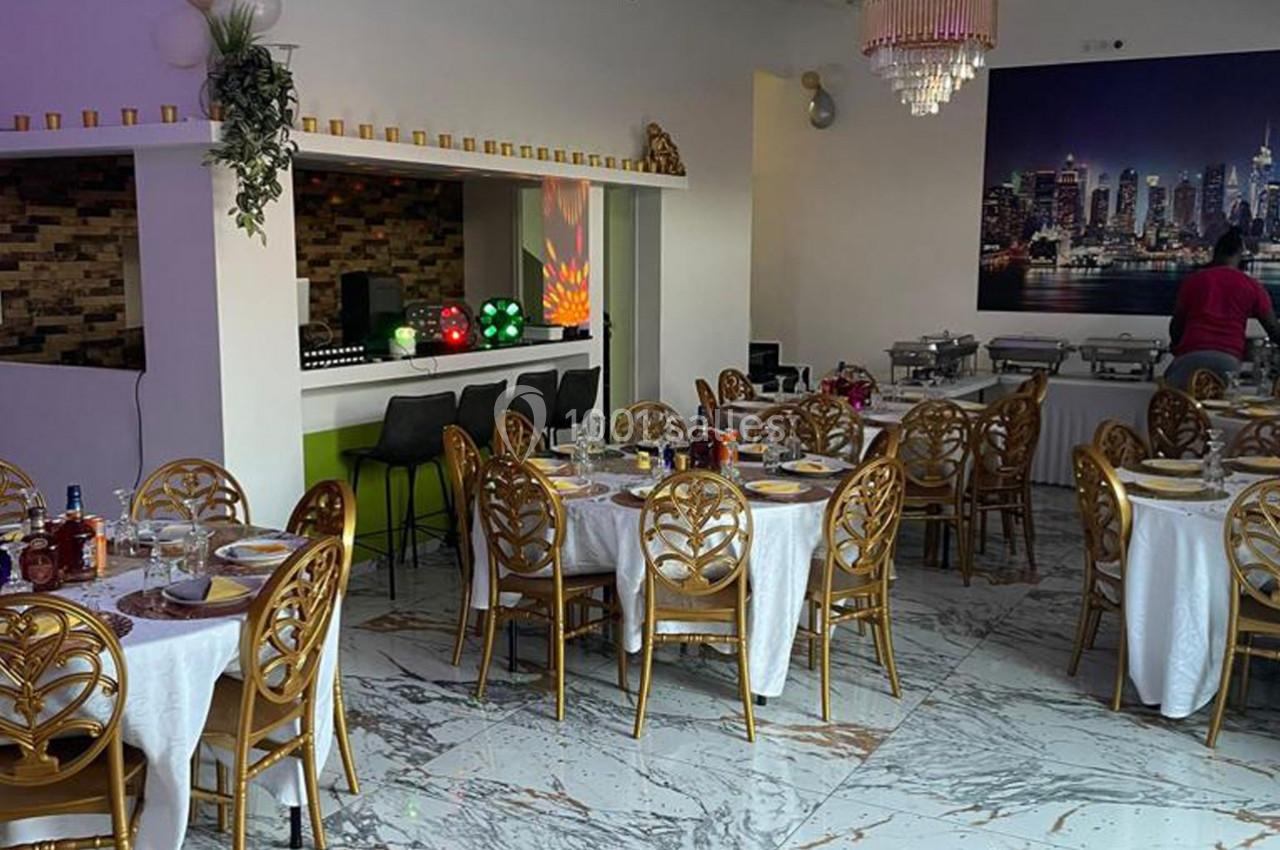 Salle de réception élégante avec tables dressées, chaises dorées, bar en arrière-plan et décoration lumineuse.