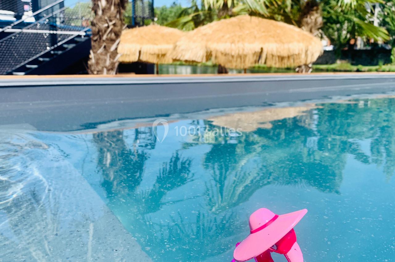 Jouet rose en forme de flamant flottant dans une piscine, avec des palmiers et une paillote en arrière-plan.