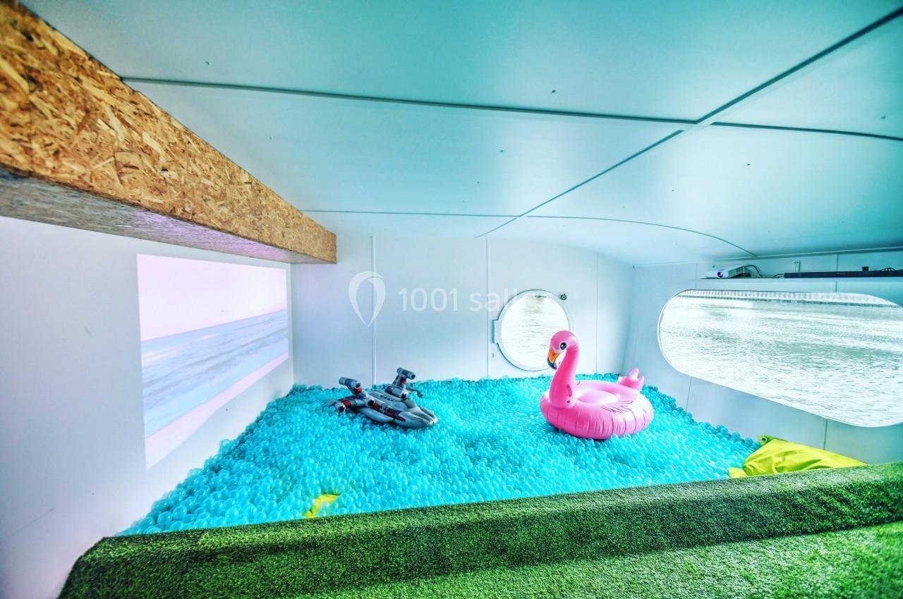 Piscine intérieure remplie de balles bleues avec une bouée rose en forme de flamant rose et vue sur l'eau par un hublot.