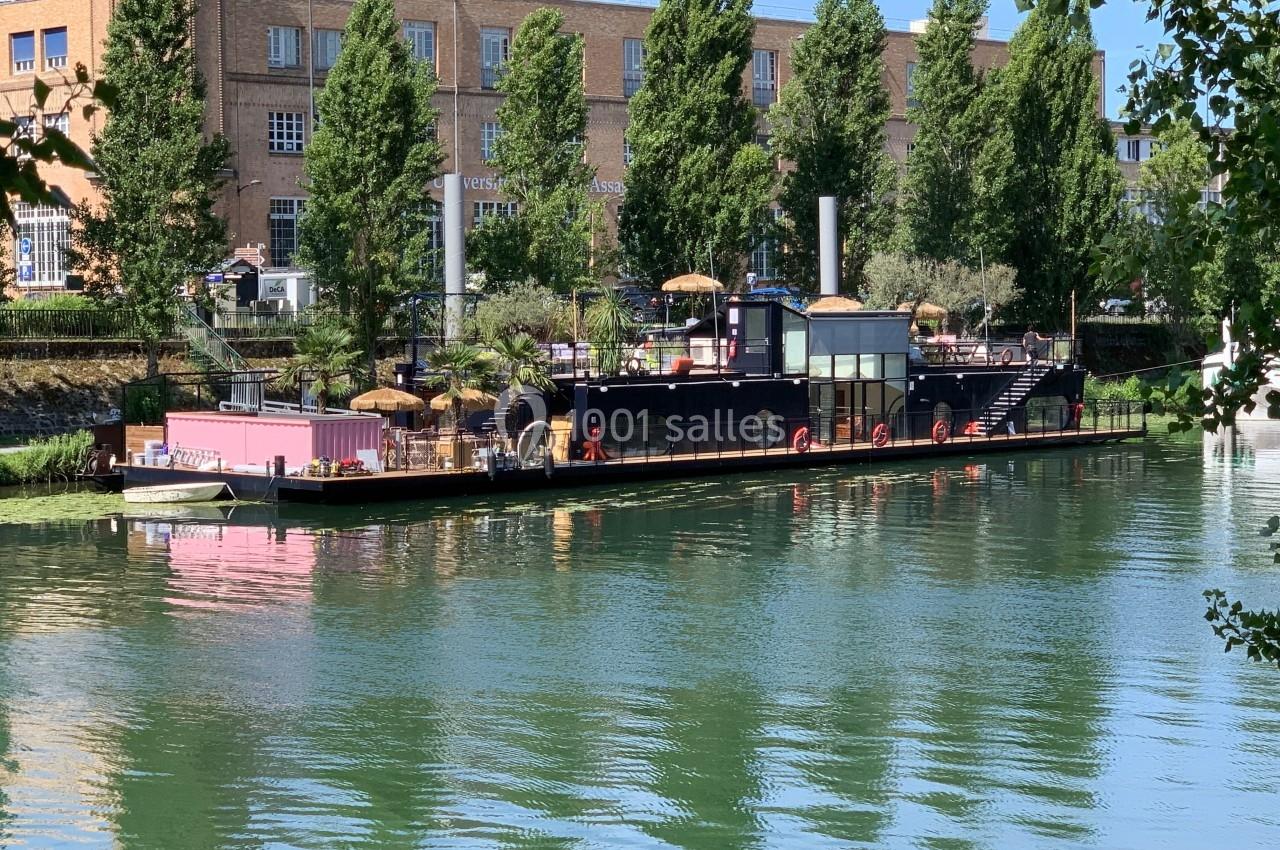 Bateau aménagé en terrasse flottante avec parasols, amarré sur une rivière bordée d'arbres et de bâtiments en briques.