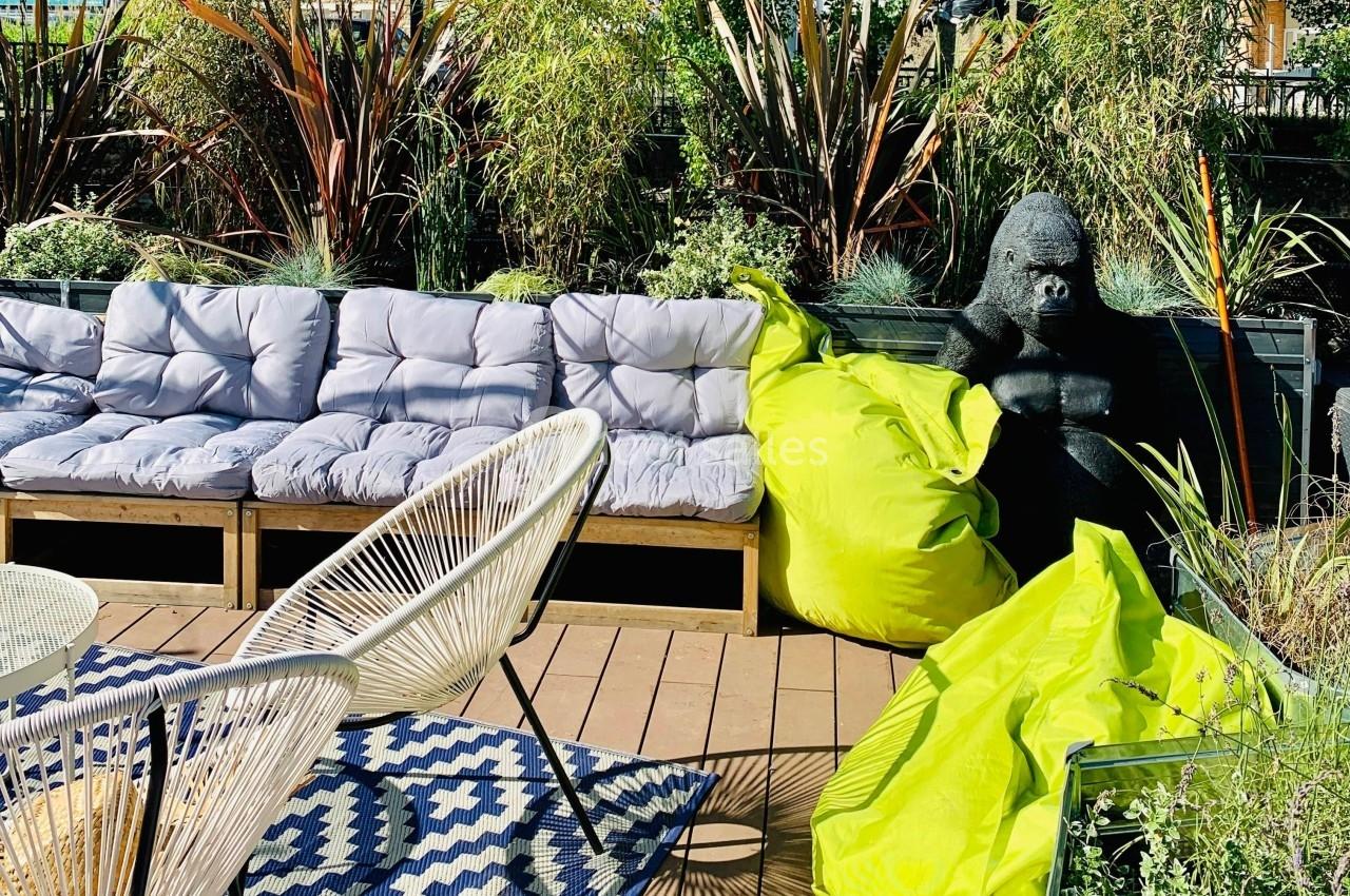 Terrasse avec canapé gris, fauteuils, poufs verts, statue de gorille noir et plantes en arrière-plan.