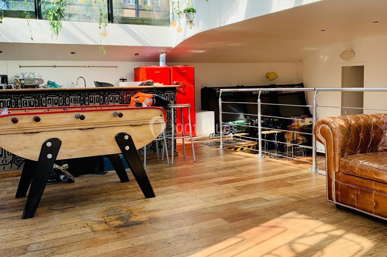 Espace lumineux avec baby-foot, canapé en cuir, cuisine ouverte et mezzanine, parquet en bois au sol.