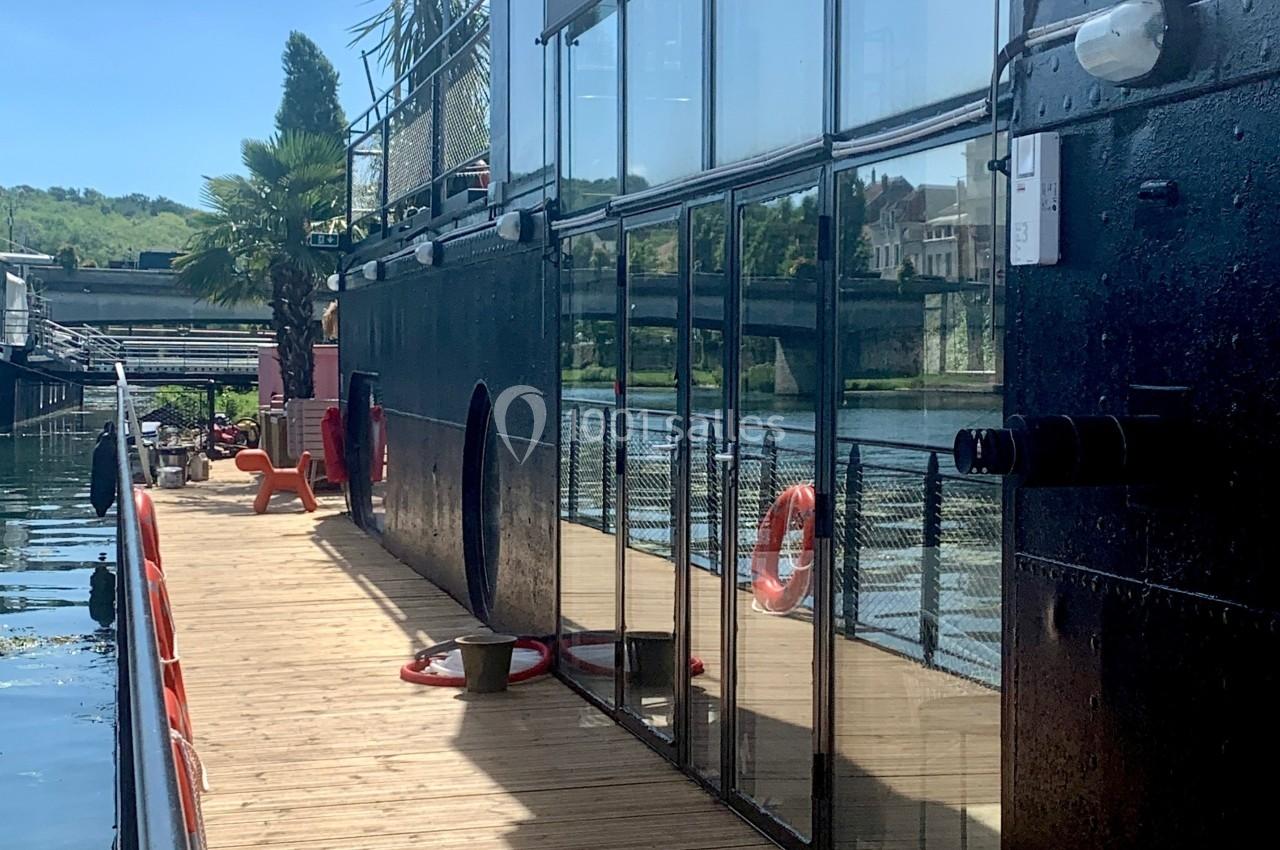 Vue latérale d'une péniche avec une passerelle en bois, des bouées de sauvetage et un paysage fluvial en arrière-plan.
