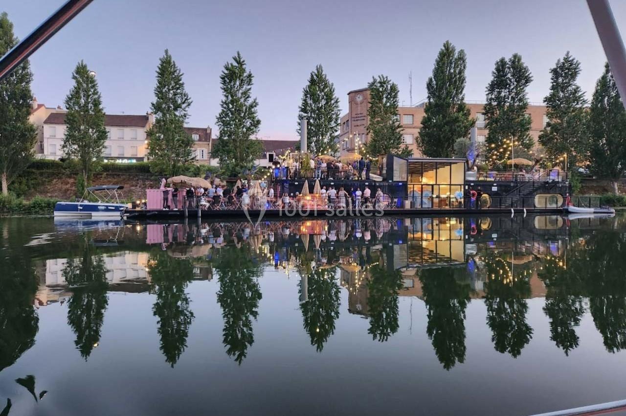 Des personnes rassemblées sur une péniche illuminée au bord d'un canal, entourée d'arbres et de bâtiments.