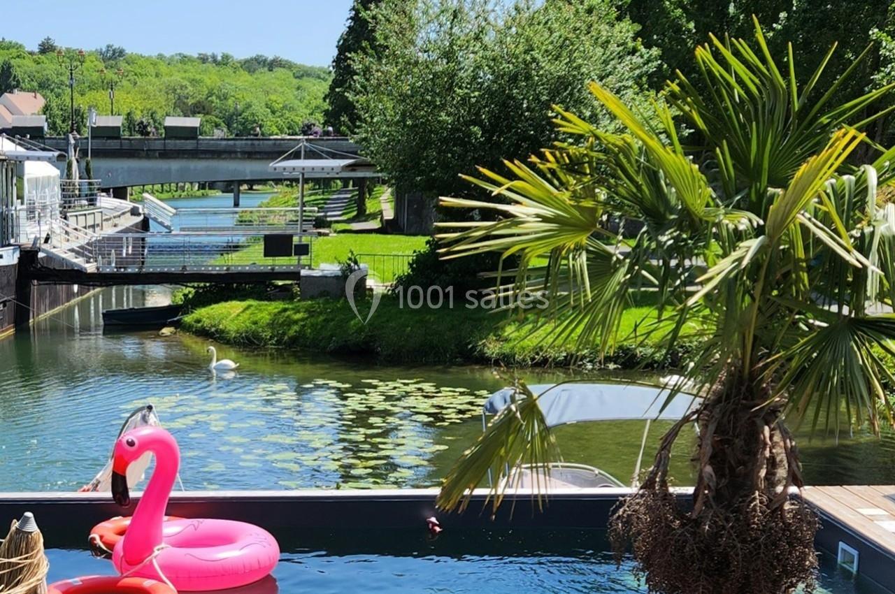 Piscine avec une bouée flamant rose en premier plan, bordée par un canal, des arbres et un cygne au loin.