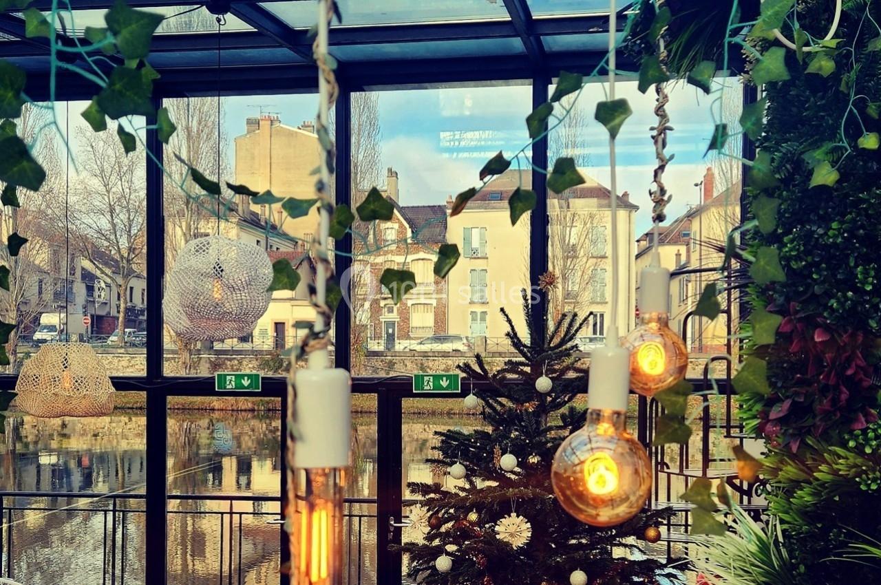 Décoration intérieure avec guirlandes lumineuses, sapin de Noël et vue sur des bâtiments à travers une verrière.