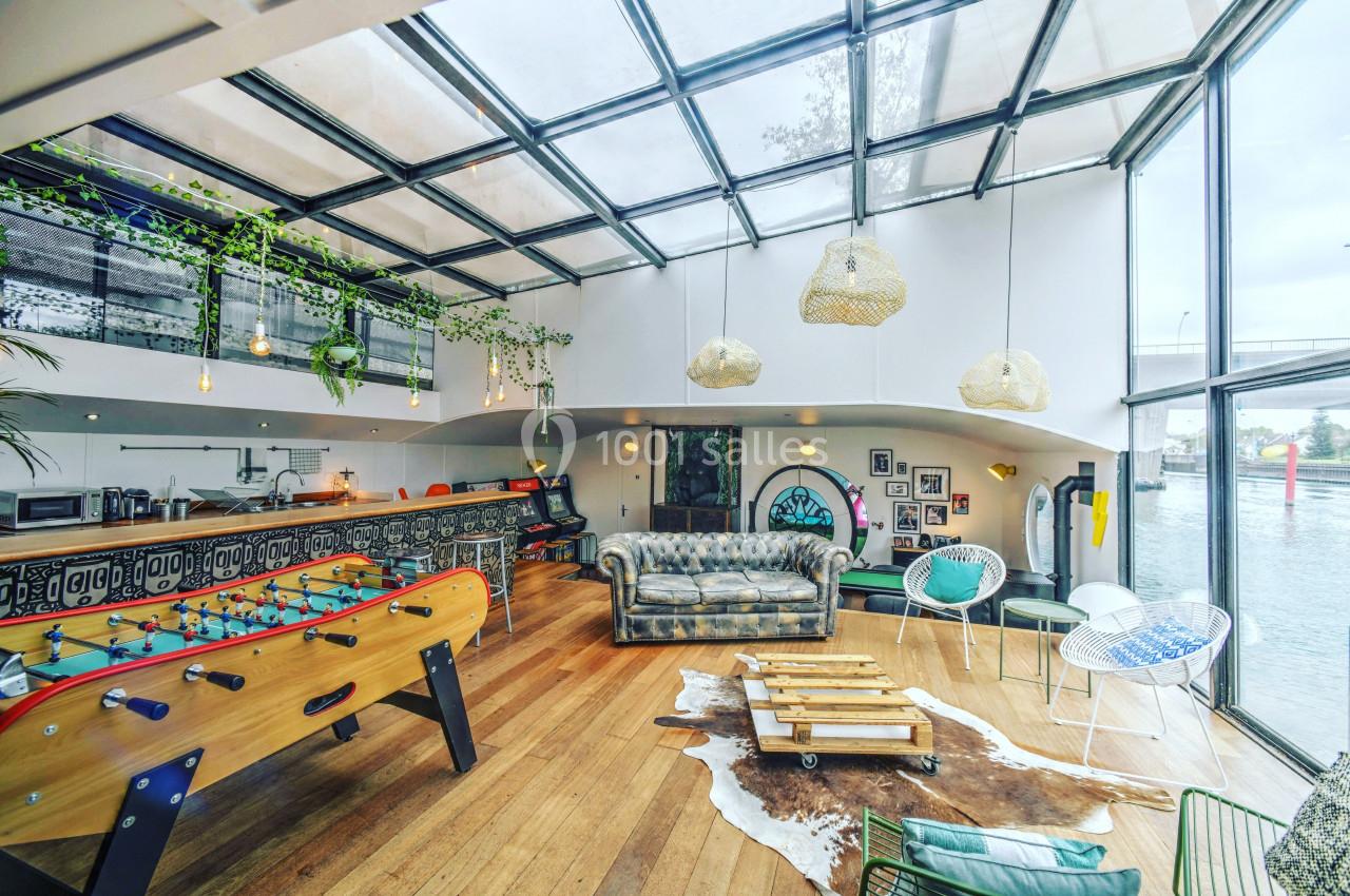 Espace lumineux avec verrière, baby-foot, canapé, décoration éclectique et vue sur un fleuve en arrière-plan.