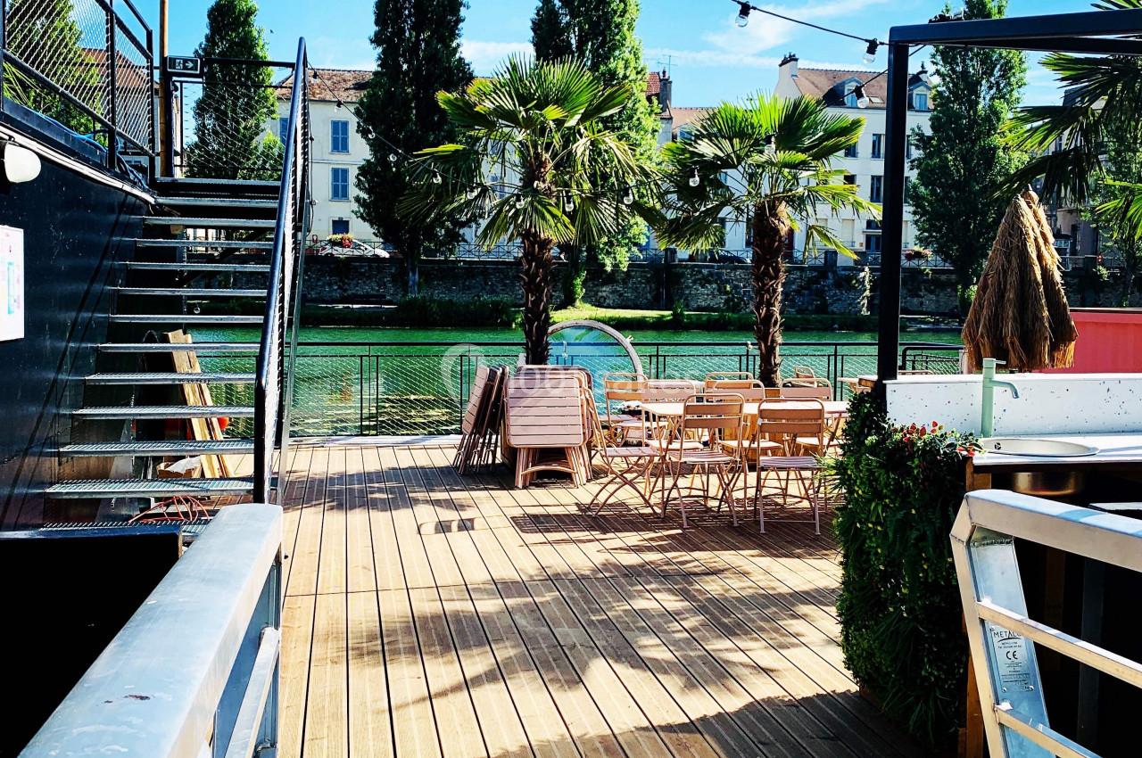 Terrasse en bois avec tables et chaises, bordée de palmiers, donnant sur un canal sous un ciel bleu.