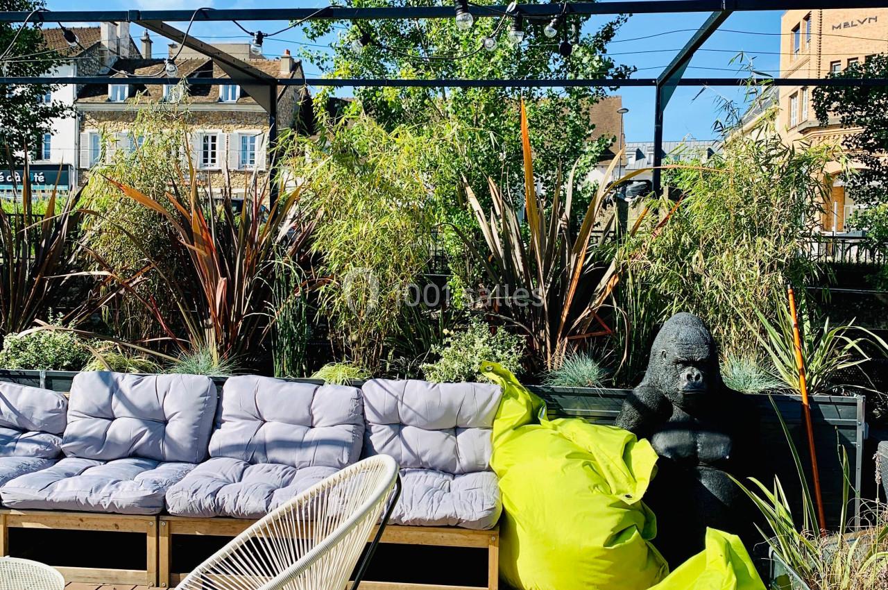 Terrasse aménagée avec canapé, fauteuils, coussins, plantes, gorille décoratif et poufs verts sous un ciel dégagé.