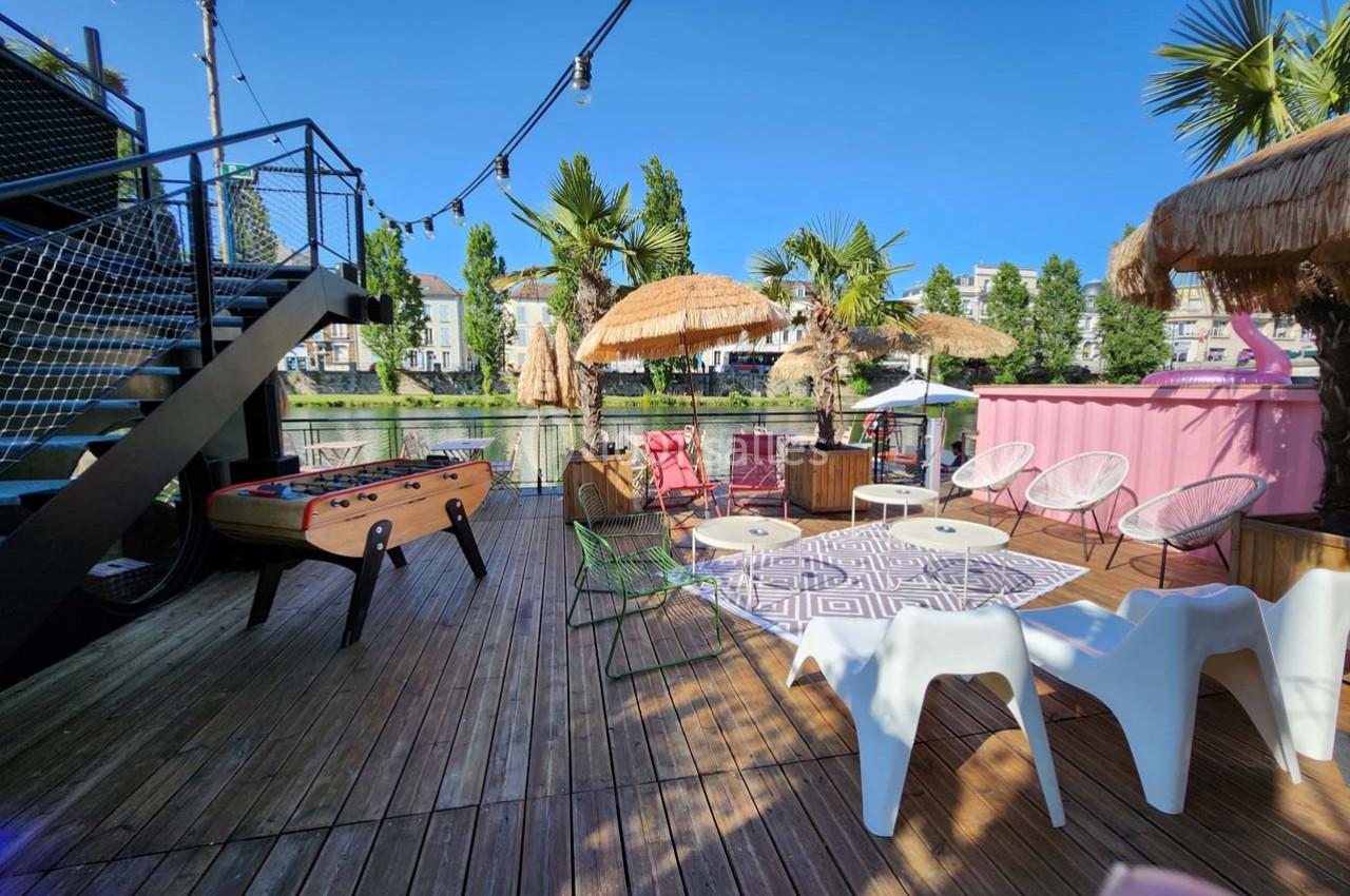 Terrasse en bois avec baby-foot, chaises, parasols en paille et vue sur un plan d'eau bordé d'arbres.