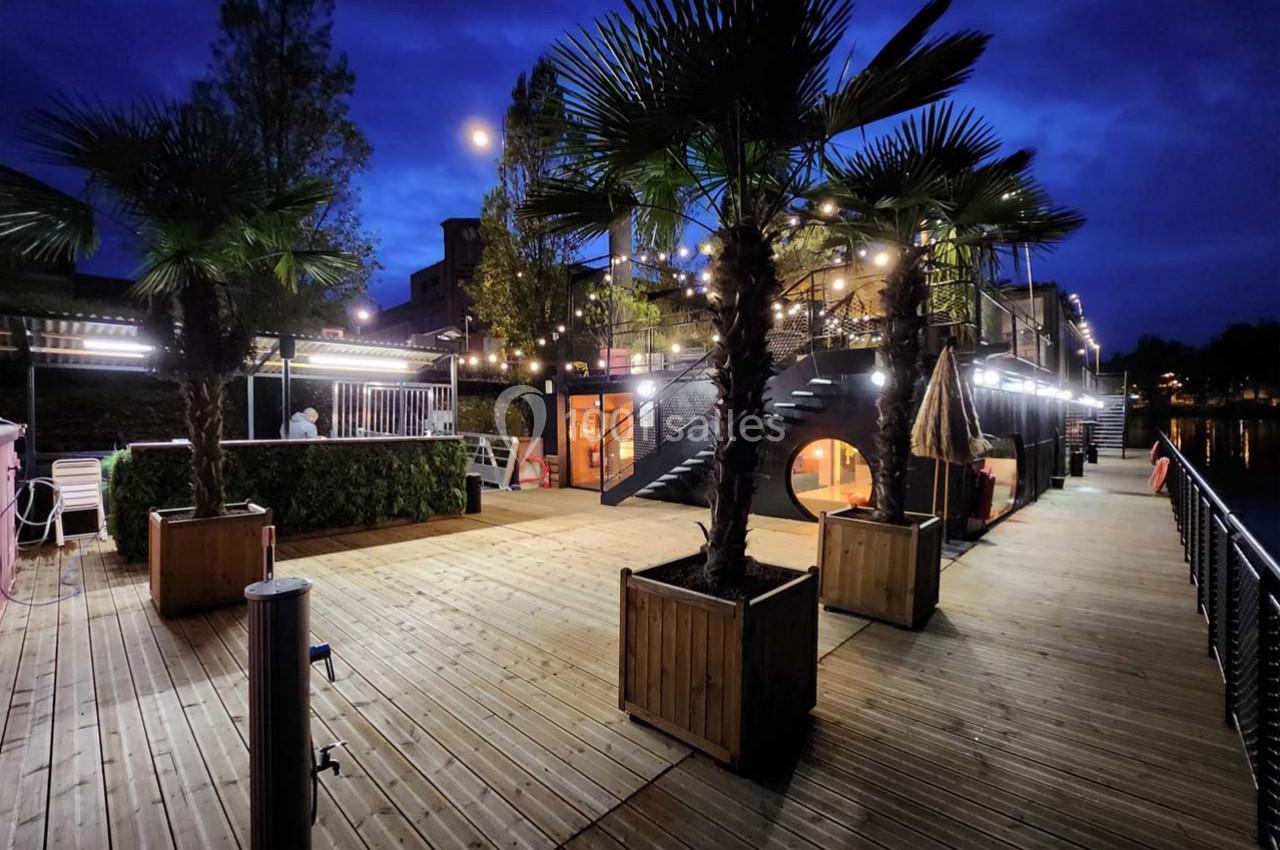 Terrasse en bois éclairée par des guirlandes lumineuses, bordée de palmiers et donnant sur un plan d'eau, de nuit.
