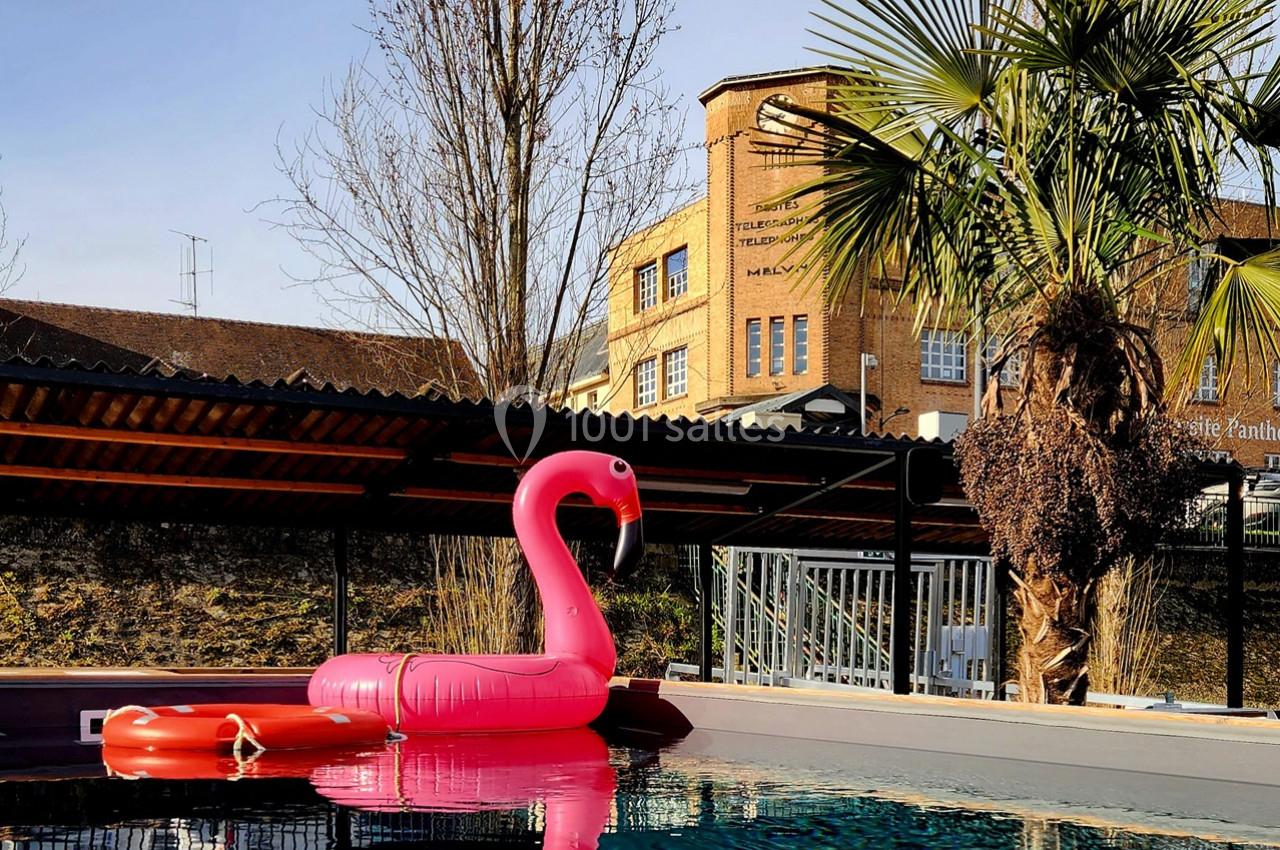 Flamant rose gonflable flottant dans une piscine extérieure, avec un palmier et des bâtiments en arrière-plan.