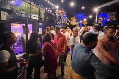 Groupe de personnes réunies dans un bar extérieur animé avec terrasse, palmiers et guirlandes lumineuses.