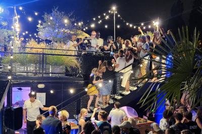 Groupe de personnes réunies dans un bar extérieur animé avec terrasse, palmiers et guirlandes lumineuses.