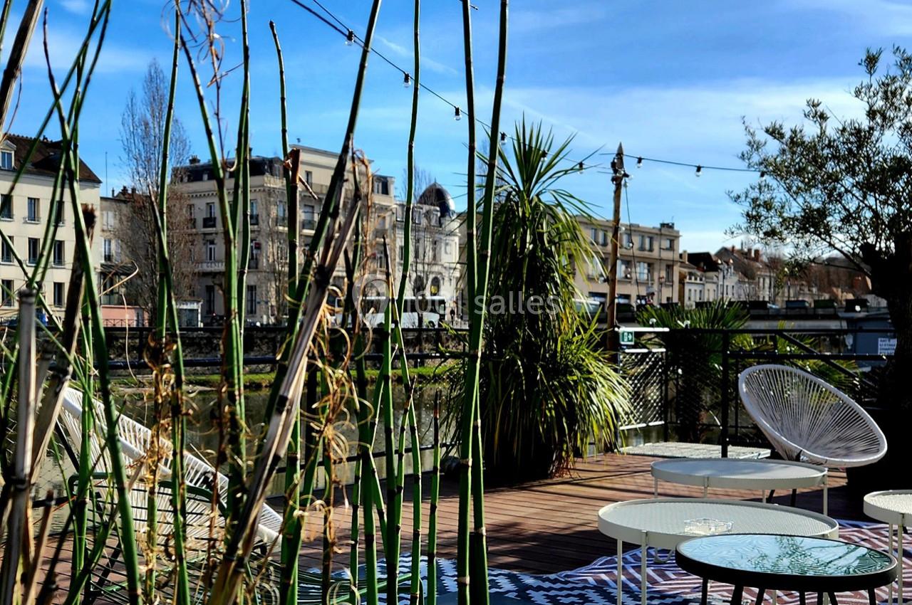 Terrasse aménagée avec des plantes, des tables et des chaises, vue sur des bâtiments et un cours d'eau en arrière-plan.