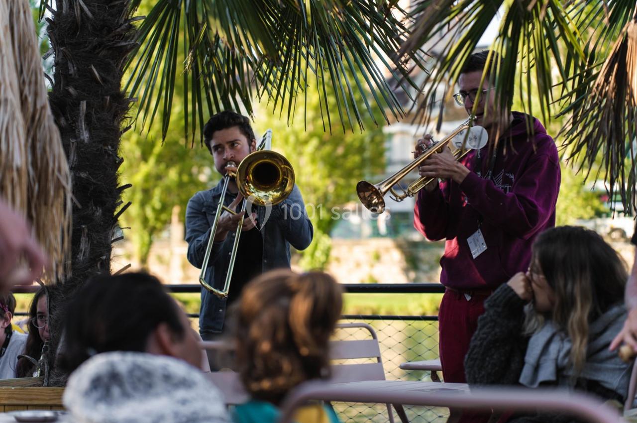 Deux musiciens jouent du trombone et de la trompette en plein air, entourés de spectateurs sous des palmiers.