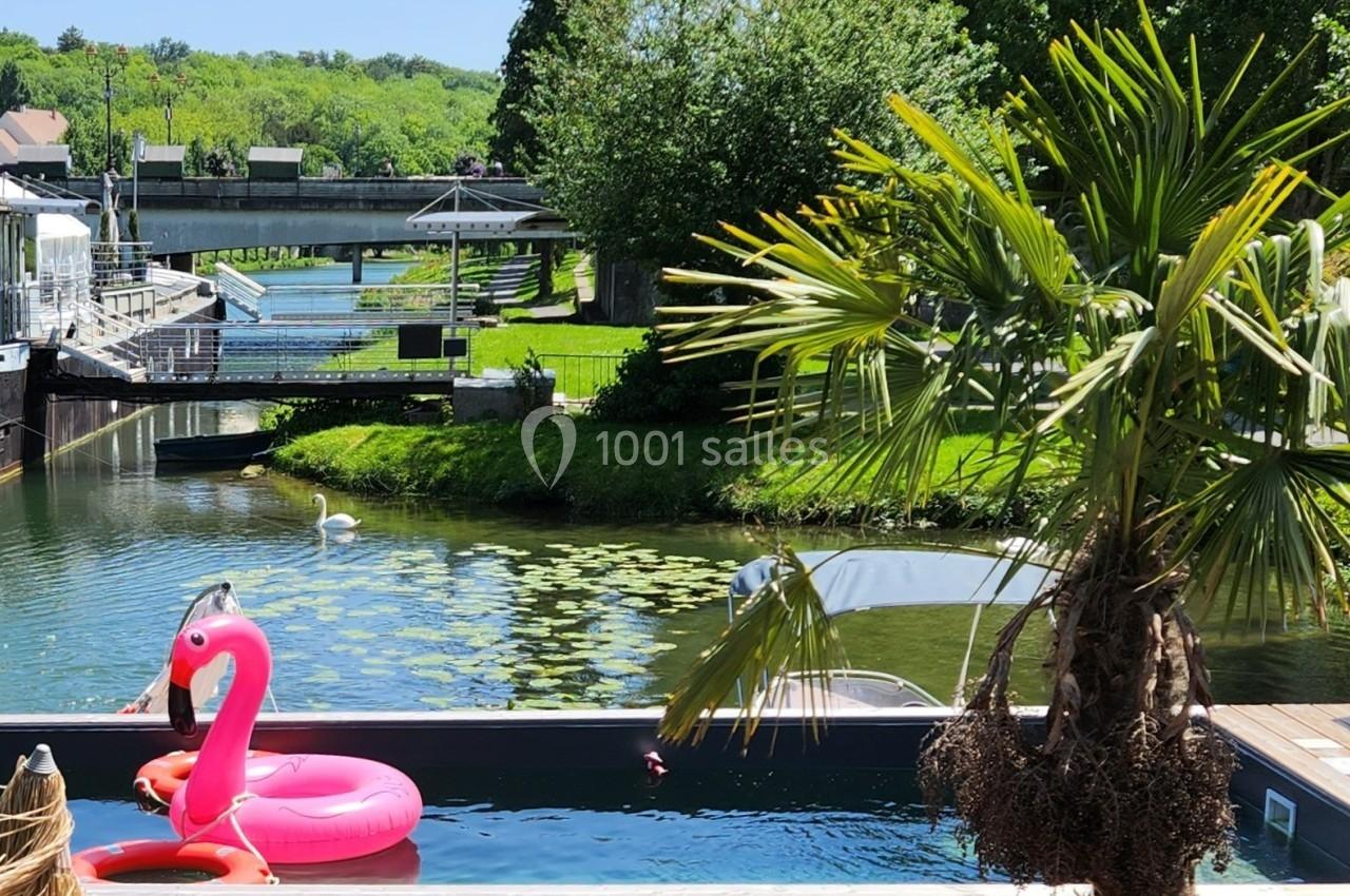 Piscine avec une bouée flamant rose, bordée d'un palmier, près d'un canal verdoyant avec un cygne et un pont.