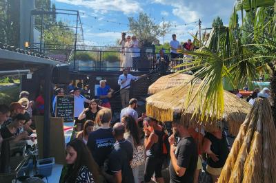 Groupe de personnes réunies dans un bar extérieur animé avec terrasse, palmiers et guirlandes lumineuses.