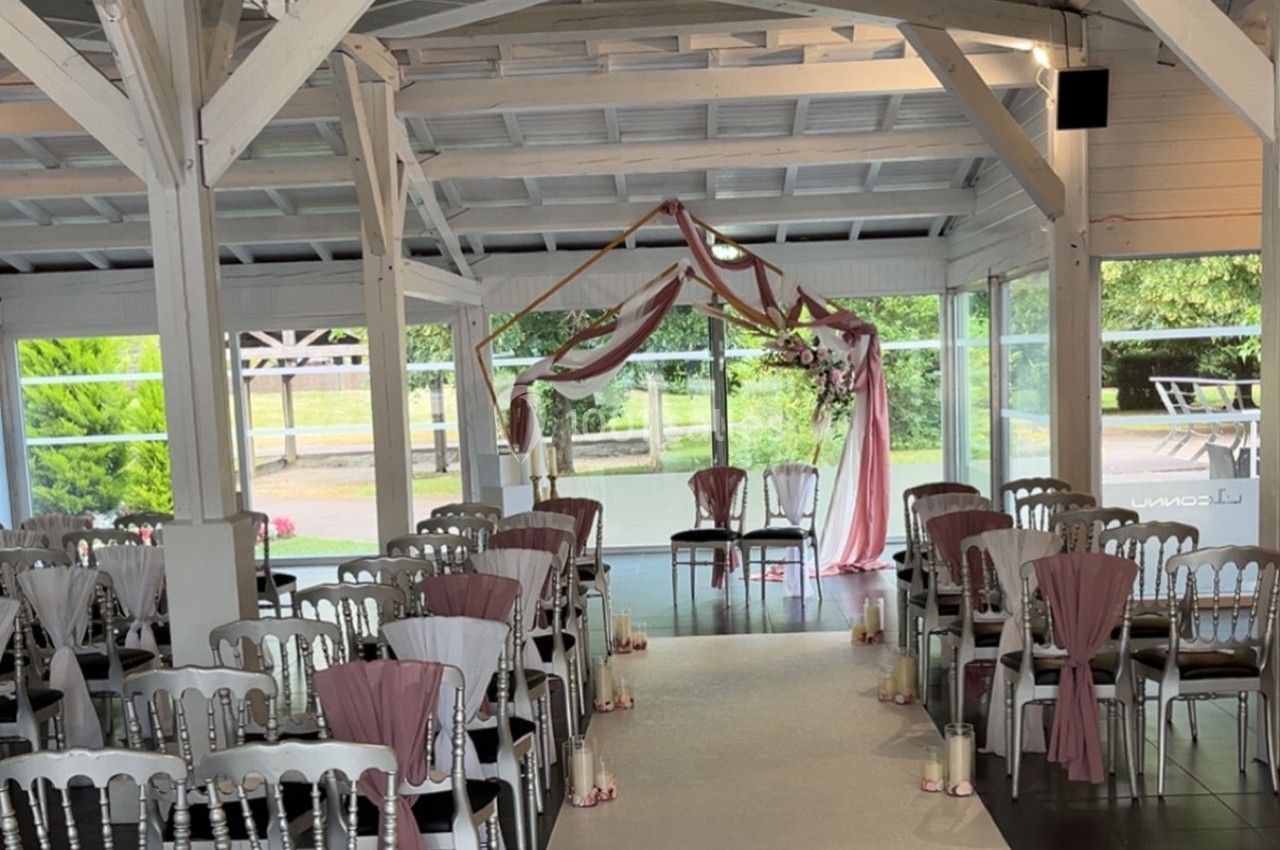 Salle de cérémonie décorée avec des chaises blanches ornées de rubans roses et une arche florale devant des baies vitrées.