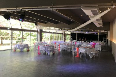 Salle de réception décorée avec des tables rondes, nappes blanches, chemins de table roses et chaises transparentes.