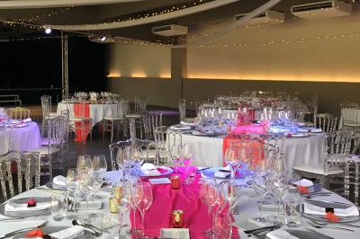 Salle de réception décorée avec des tables rondes, nappes blanches, chemins de table roses et chaises transparentes.