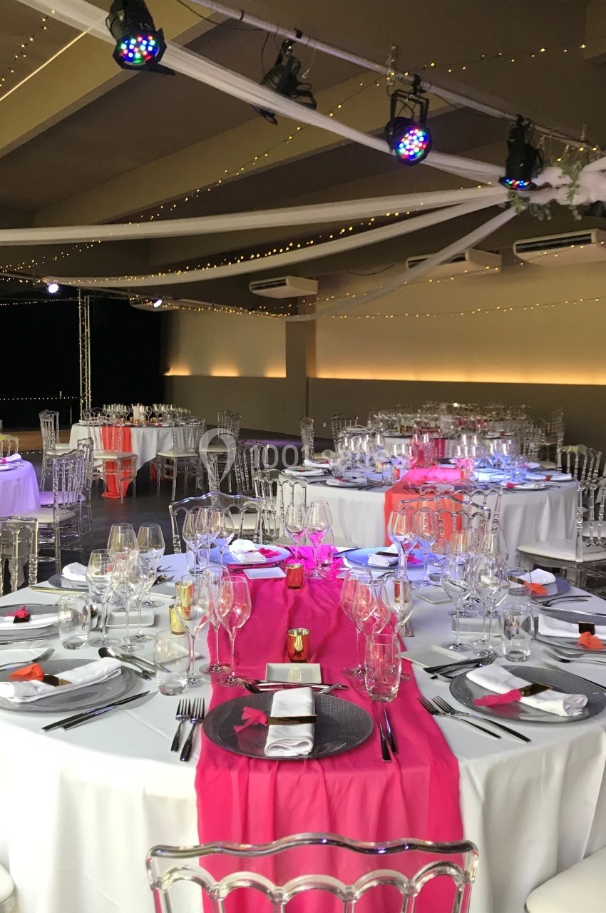 Salle de réception décorée avec des tables rondes, nappes blanches, chemins de table roses et chaises transparentes.