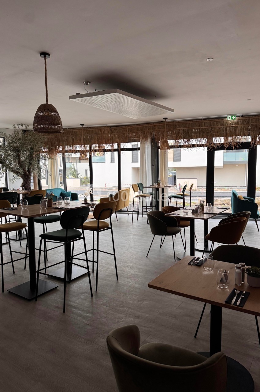 Salle de restaurant moderne et lumineuse avec des tables dressées, des chaises colorées et de grandes baies vitrées.