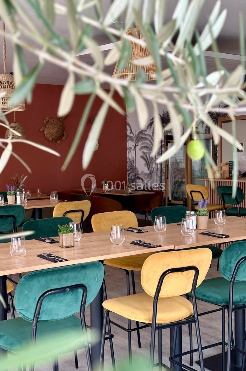 Salle de restaurant lumineuse avec des tables en bois, chaises vertes et jaunes, et décoration végétale et murale.