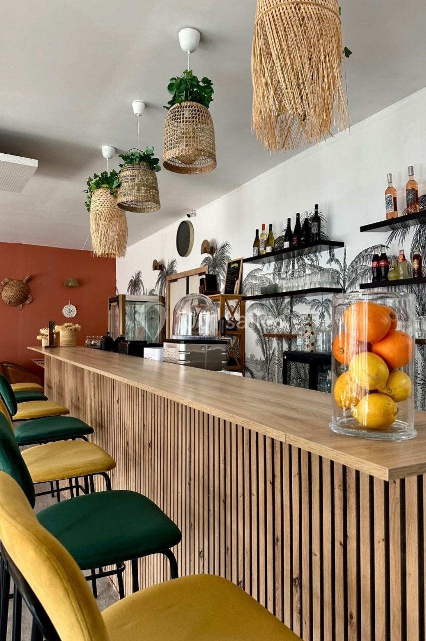 Comptoir en bois avec tabourets colorés, suspensions en osier, étagères garnies de bouteilles et corbeille de fruits.