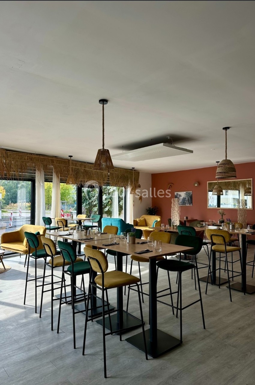 Salle lumineuse avec tables hautes, chaises colorées, grandes fenêtres et décorations en fibres naturelles.