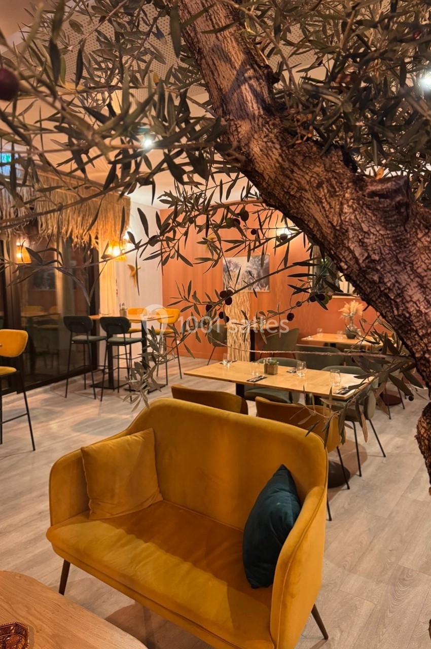 Intérieur d'un restaurant avec un canapé jaune, des tables en bois, des chaises hautes et un arbre décoratif au premier plan.