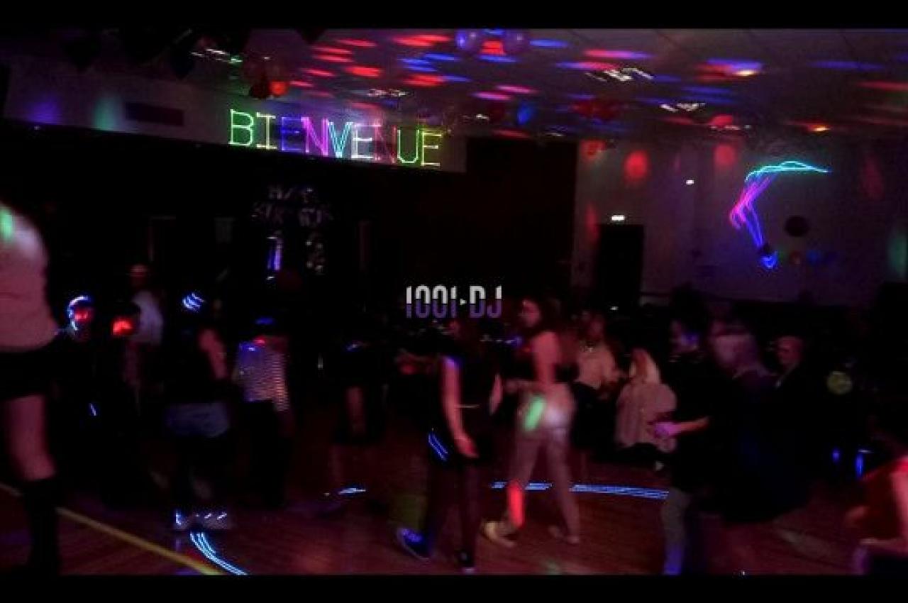 Personnes dansant dans une salle sombre éclairée par des lumières colorées, avec un panneau ’Bienvenue’ lumineux.