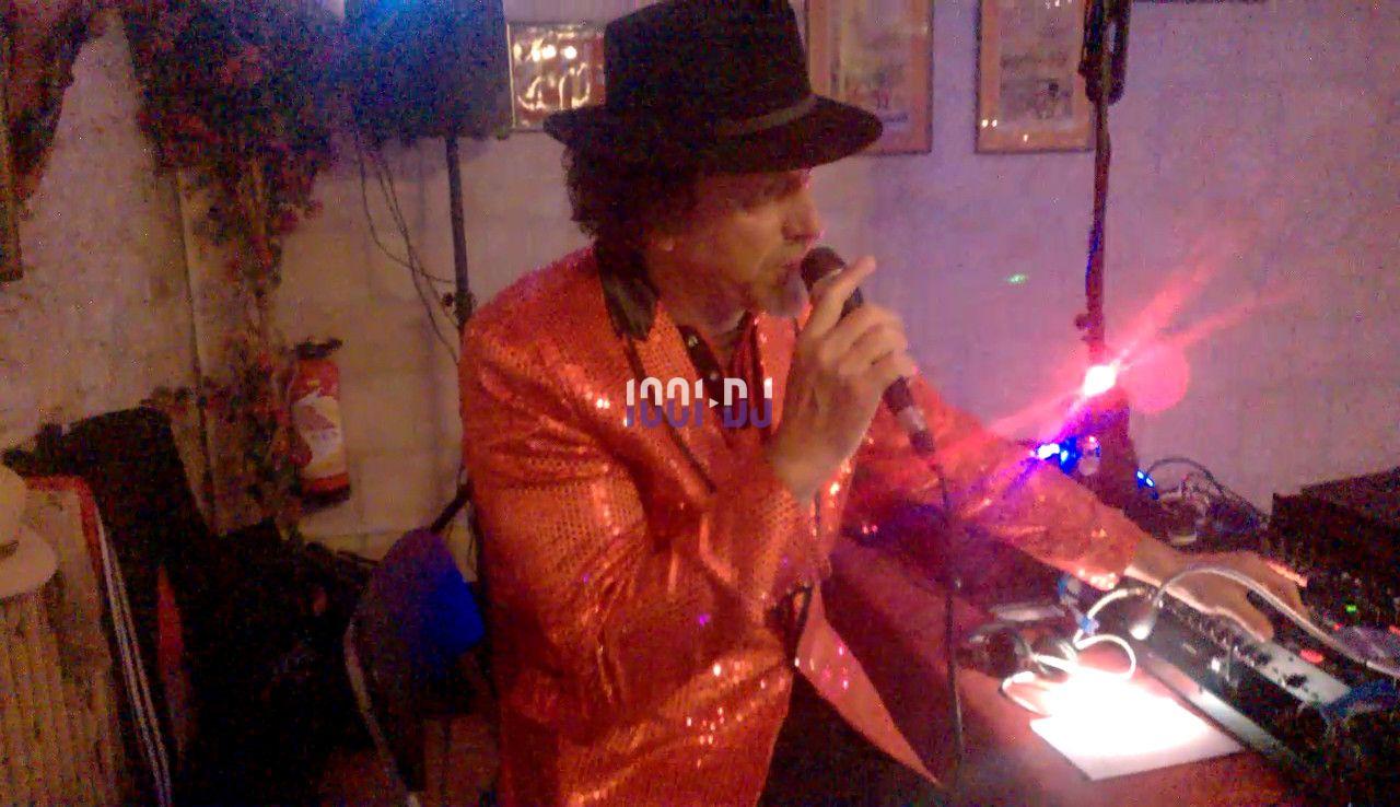 Un chanteur en veste rouge scintillante et chapeau noir utilise un micro lors d'une performance en intérieur.