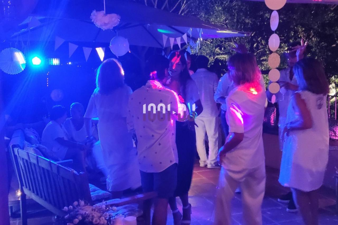 Des personnes en tenue blanche dansent et discutent sous des lumières colorées lors d'une soirée en extérieur.