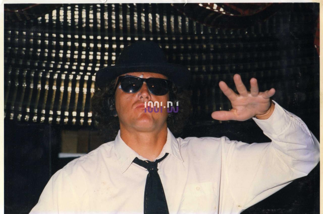 Homme portant des lunettes de soleil, un chapeau noir et une cravate, levant une main devant un fond sombre.