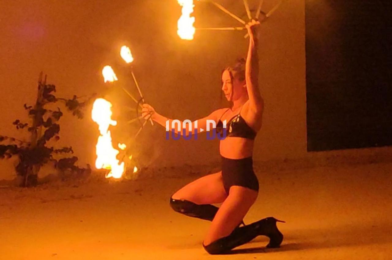 Une femme en tenue noire manipule des éventails enflammés, agenouillée dans un espace extérieur éclairé par les flammes.