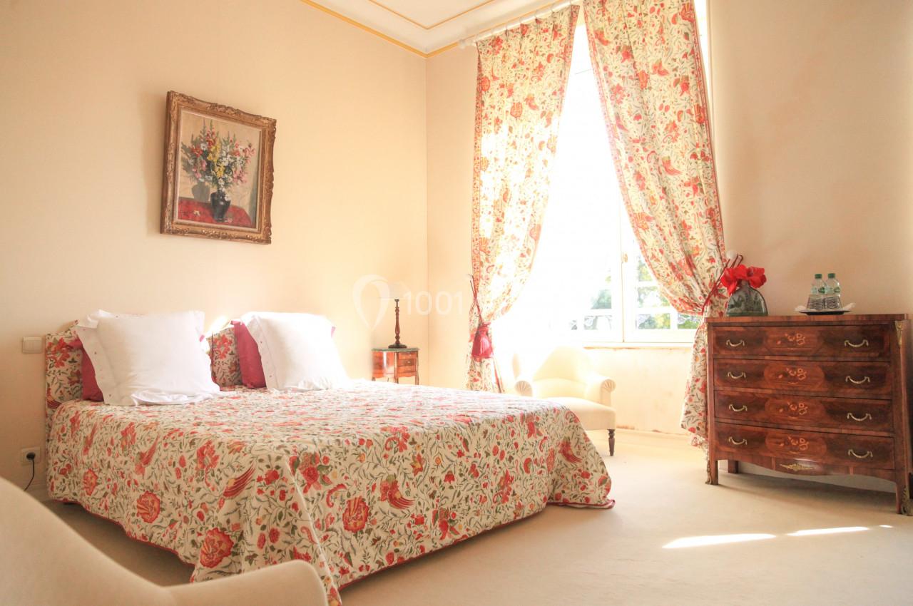 Chambre lumineuse avec lit double, couvre-lit fleuri, commode en bois, fauteuil et grande fenêtre avec rideaux assortis.