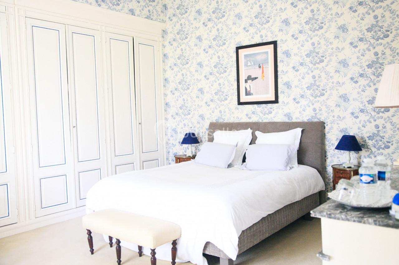 Chambre lumineuse avec lit double, murs à motif floral bleu, armoire blanche et tables de chevet avec lampes bleues.