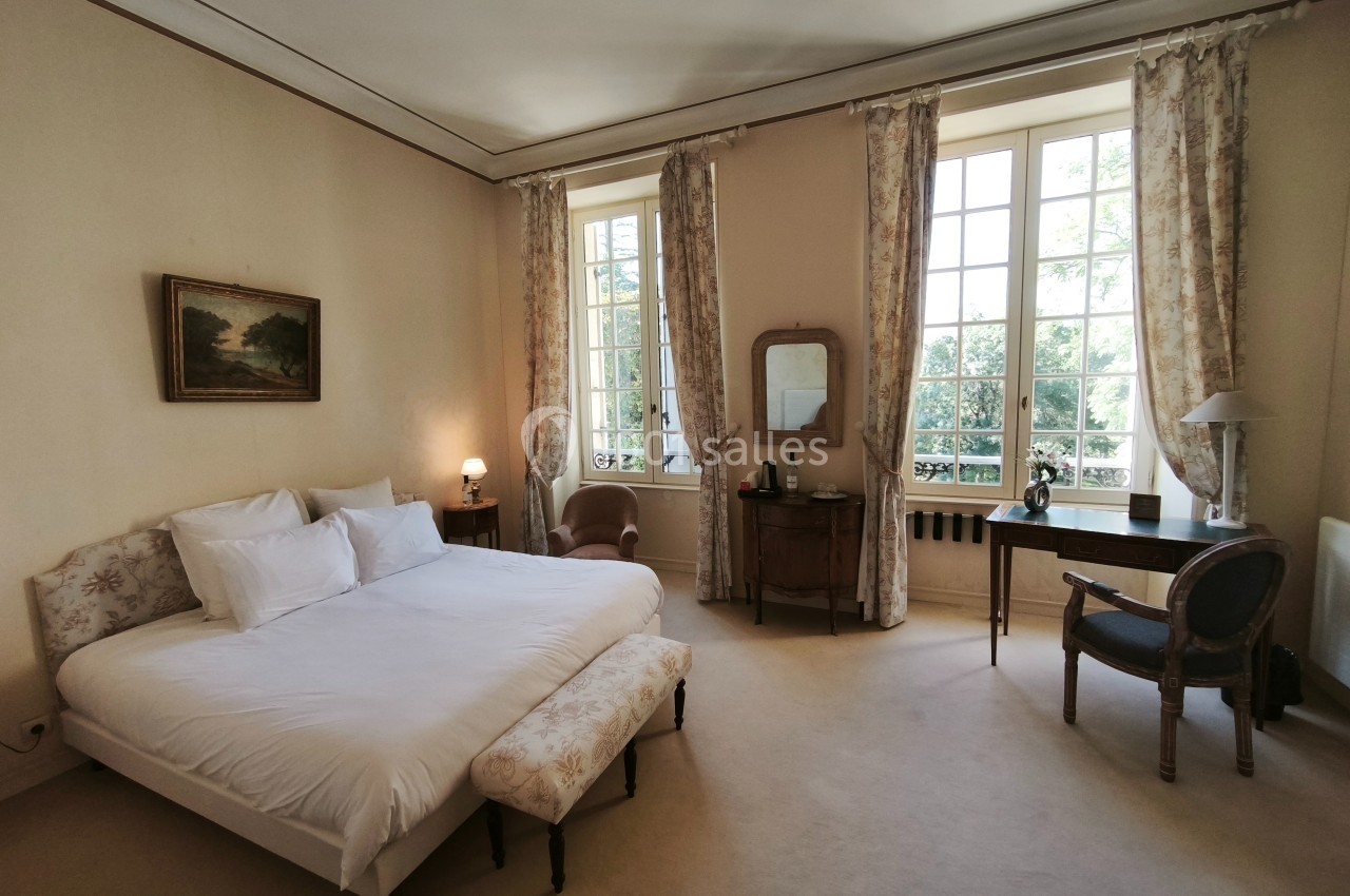 Chambre lumineuse avec un lit double, des meubles anciens, de grandes fenêtres et des rideaux clairs.