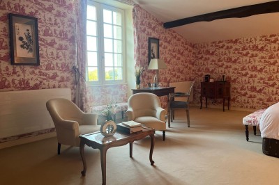 Chambre lumineuse avec murs en toile de Jouy rouge, mobilier classique, table basse et fauteuils près d'une fenêtre.