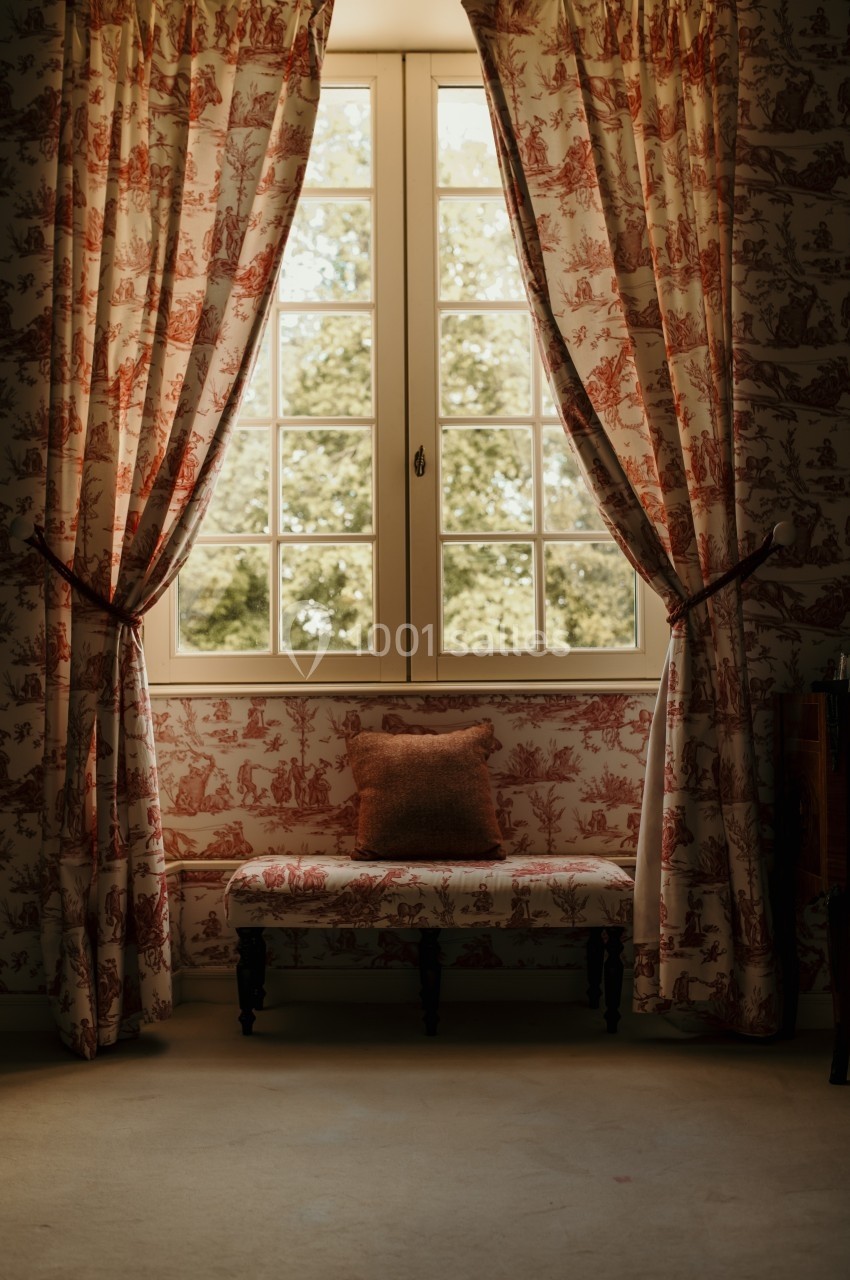 Fenêtre encadrée de rideaux à motifs rouges, avec un banc assorti et un coussin, éclairée par la lumière naturelle.