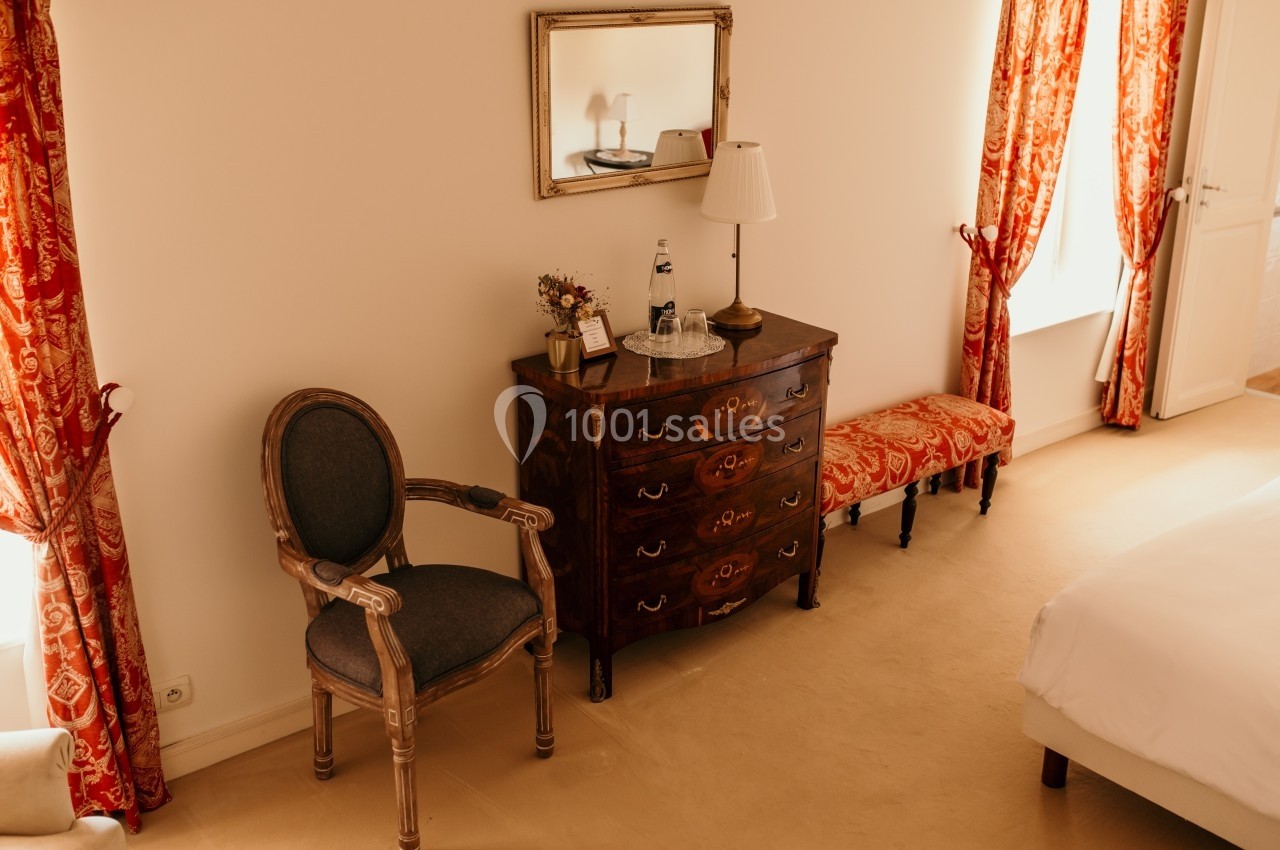 Chambre lumineuse avec commode en bois, chaise, miroir, rideaux rouges et lit partiellement visible.