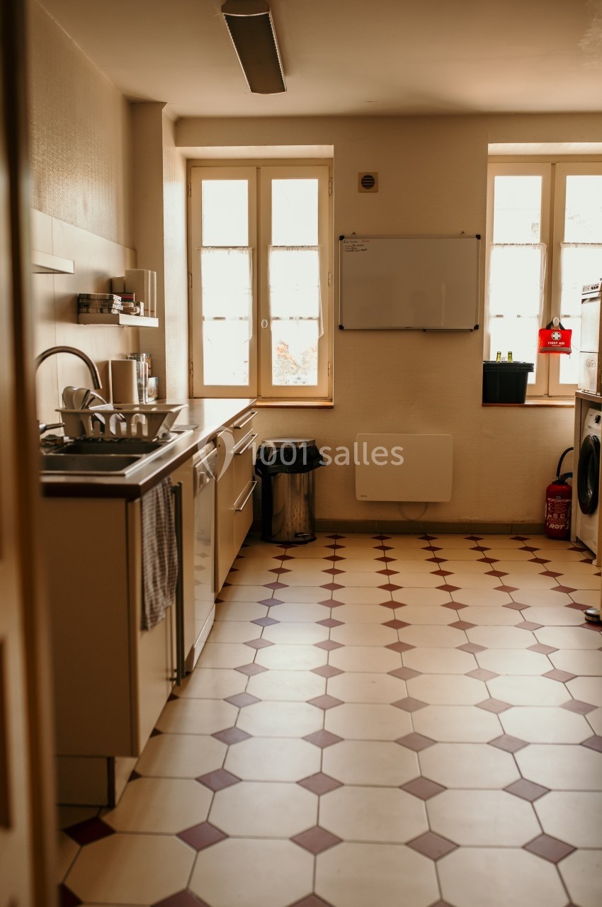 Cuisine lumineuse avec évier, lave-vaisselle, poubelle et sol en carrelage beige et rouge, éclairée par deux fenêtres.