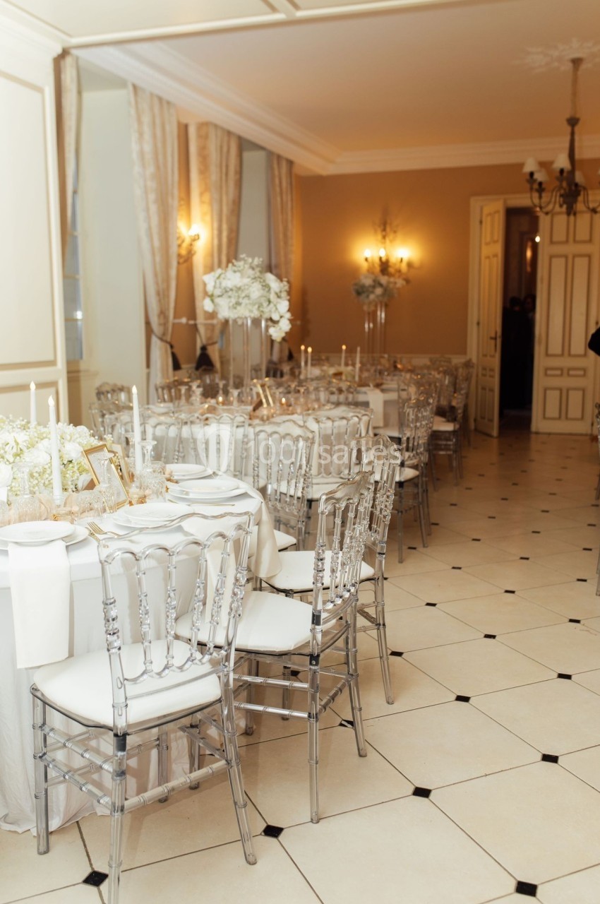 Salle élégante avec tables rondes dressées, chaises transparentes et décoration florale blanche dans un cadre lumineux.