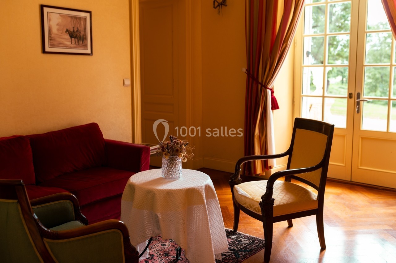 Salon lumineux avec canapé rouge, deux fauteuils, table ronde avec nappe blanche et vase, près d'une porte-fenêtre.