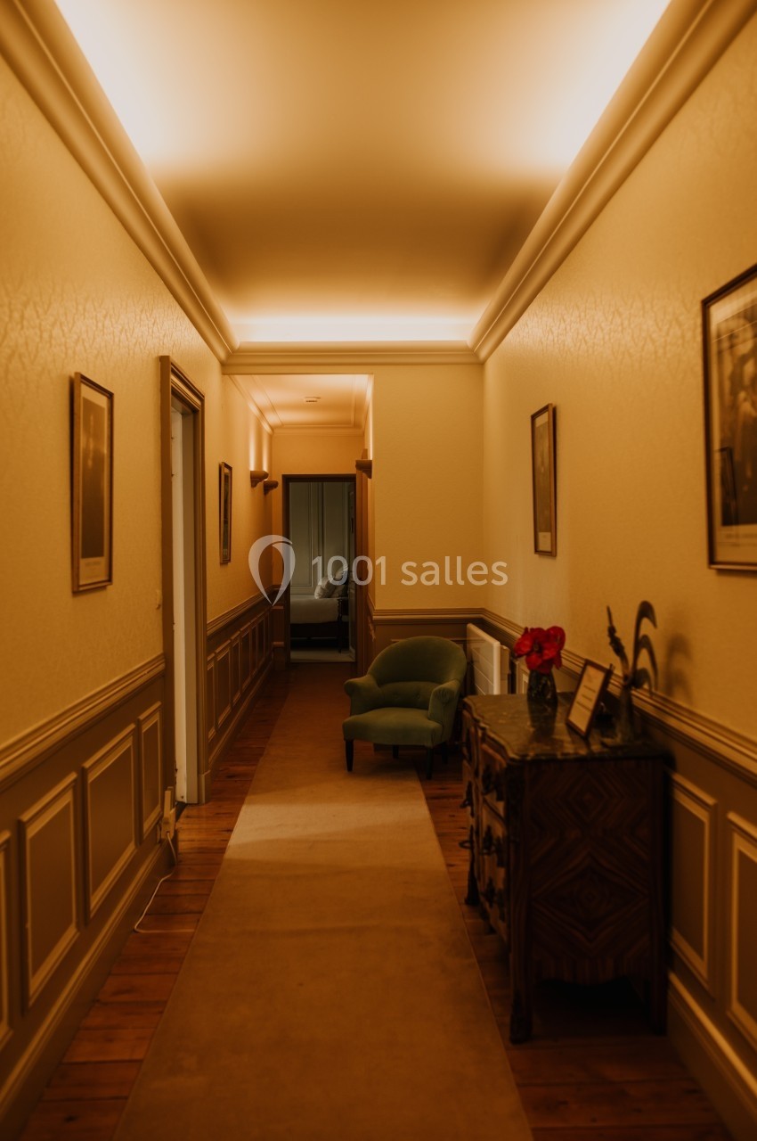 Couloir étroit avec éclairage chaleureux, tapis beige, fauteuil vert, commode en bois et cadres accrochés aux murs.