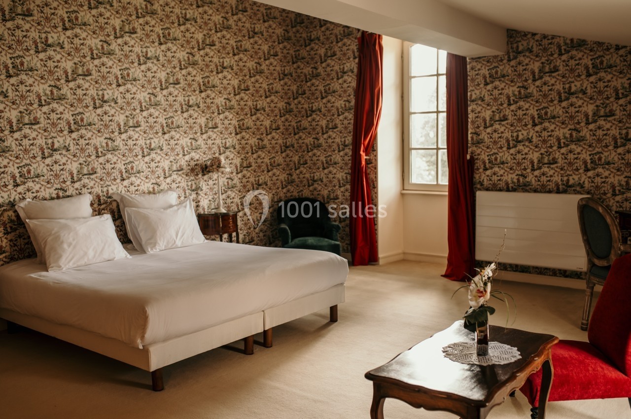 Chambre lumineuse avec lit double, fauteuils, table basse, papier peint fleuri et rideaux rouges encadrant une fenêtre.