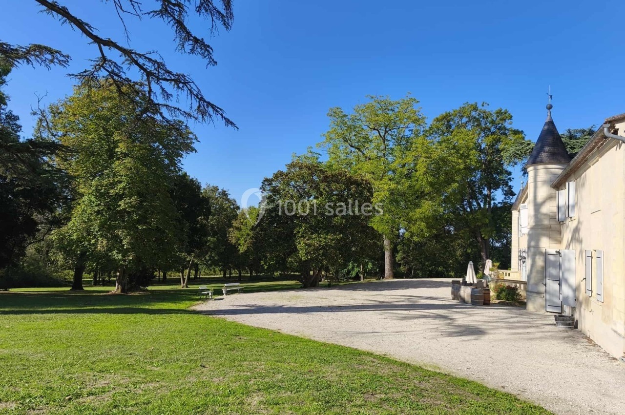 Allée gravillonnée bordée d'arbres menant à un bâtiment ancien avec une tourelle, par une journée ensoleillée.