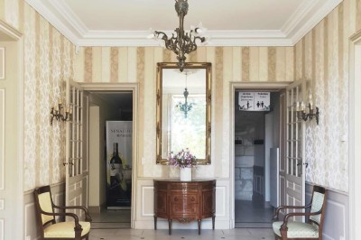 Façade claire avec volets blancs, lanterne en fer forgé et jardinières de fleurs roses en premier plan.