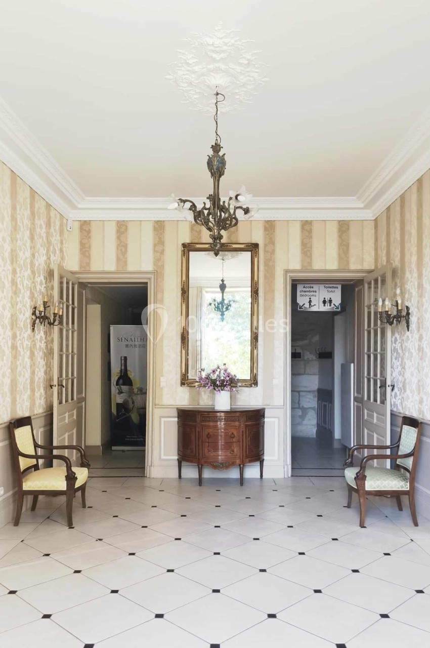 Entrée lumineuse avec sol carrelé, meubles anciens, miroir doré, lustre central et portes ouvertes sur d'autres pièces.